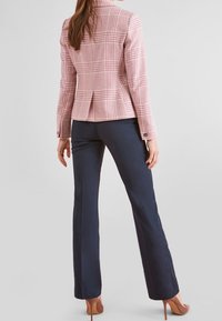 Blazer en tartan rose avec une coupe ajustée, col cranté et accents en bouton, associé à un pantalon marine et des talons marron clair.