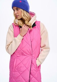 Różowa pikowana kamizelka puchowa z kapturem, z czarnymi wykończeniami, kieszeniami i błyszczącym wykończeniem. Noszona na kremowej bluzie i niebieskiej czapce.