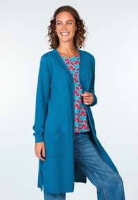 Teal gestrickte Strickjacke mit strukturiertem Muster, offener Front und Seitentaschen, über einem floral gemusterten Oberteil in Rot und Türkis getragen.