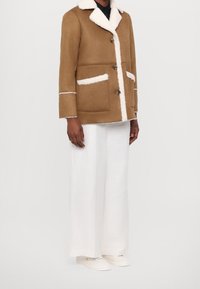 Hnědá umělá sukně s bílým límcem ze syntetické kožešiny, dvěma předními kapsami a bílým lemem. Nosit s bílými širokými kalhotami a teniskami.