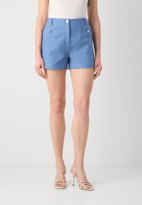 Pantalones cortos tejidos en azul con cintura alta, parte delantera plana, dos botones decorativos y bolsillos laterales. Combinados con sandalias de tacón alto de tiras en color beige.