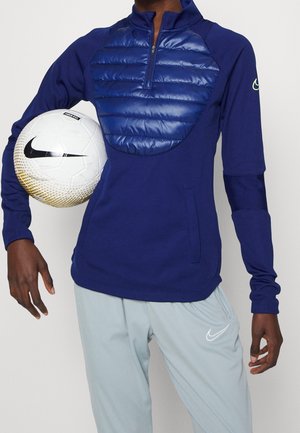 Personne portant un pull quart-zip bleu marine et un pantalon Nike gris clair tient un ballon de football Nike blanc sous le bras.