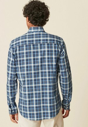 Blau kariertes Hemd mit langen Ärmeln und geknöpften Manschetten, mit weißen und dunkleren blauen Linien. Leicht abgerundeter Saum und Rückenpasse-Detail.