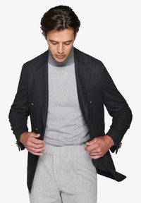 Sort dobbeltradet blazer i struktureret stof, grå mønstret turtleneck og lysegrå skræddersyede bukser. Modellen justerer blazeren.