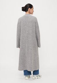 Lang grå strikket cardigan med en avslappet passform. Har nedfalte skuldre, ribbestrikkede mansjetter og en jevn tekstur. Bært over blå jeans og sneakers.