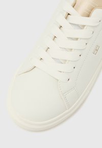Zapatilla deportiva de cuero blanco con puntera redonda, cordones planos y sutiles detalles dorados en la lengüeta. Textura suave con costuras mínimas.