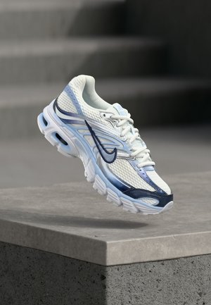 Biały i jasnoniebieski but sportowy z ciemnoniebieskim logo Swoosh unoszący się nad szarym betonowym blokiem.