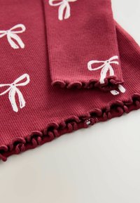 Burgundská ribbovaná tkanina s bielymi vzormi mašlí; obsahuje volánové okraje na manžetách a dolnom leme pre pridanie textúry a detailov.