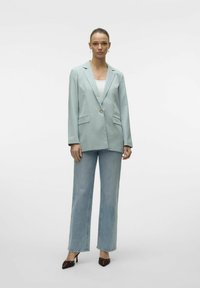Blazer verde chiaro con un singolo bottone dorato, rever a punta e tasche frontali, abbinato a jeans azzurri a vita alta e tacchi appuntiti.