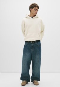 Helle beigen Oversized-Hoodie mit einer Fronttasche, kombiniert mit weit geschnittenen blauen Jeans und beigen Sneakers. Einfaches Design und weiche Textur.