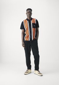 Camisa de manga corta con cuello, de rayas verticales en naranja, negro y blanco, combinada con pantalones de pana negros y zapatillas beige.