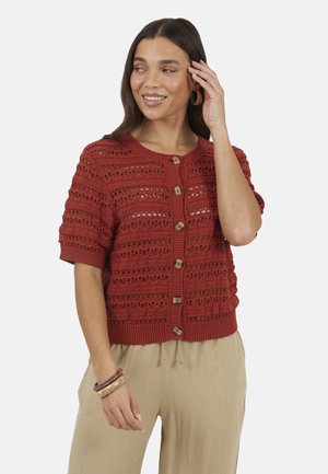 Vrouw met een roestkleurig gebreid kort mouwtje vest met knopen, beige broek en bruine armbanden, glimlachend en haar haar aanrakend.