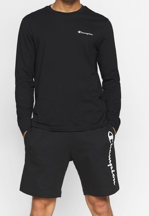 Homme portant un t-shirt noir à manches longues et un short noir avec des logos Champion blancs sur la poitrine et la jambe gauche, mains dans les poches.
