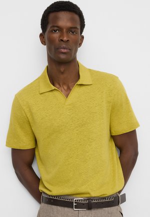 Homme portant un polo jaune à manches courtes rentré dans un pantalon beige avec une ceinture noire, debout devant un fond clair uni.