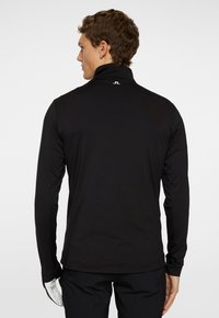 Musta pitkähihaisen polo-paita korkealla kauluksella, valmistettu sileästä kankaasta. Takana on pieni logo ja istuva muotoilu.