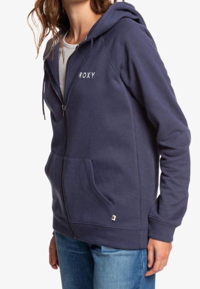 Femme portant un sweat à capuche bleu marine zippé avec le texte "ROXY" sur la poitrine et un jean bleu clair.