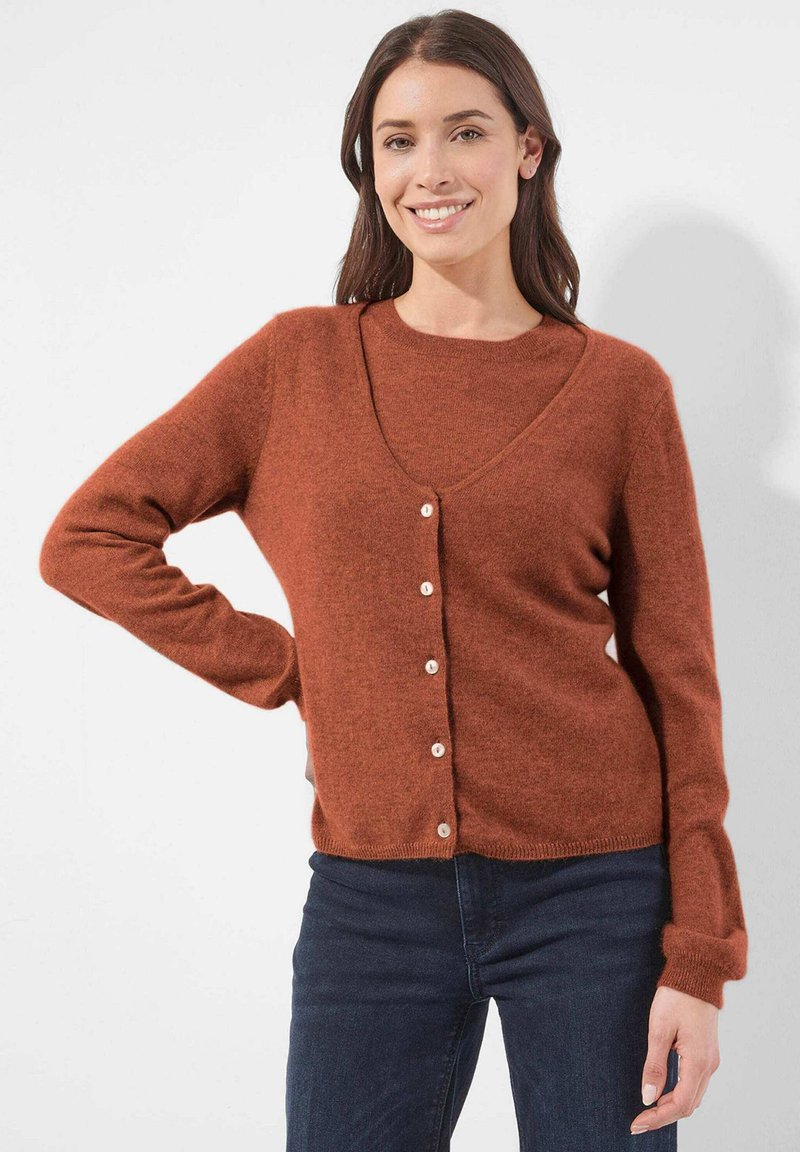 Vrouw die glimlacht, gekleed in een rustkleurige cardigan met knopen over een bijpassend T-shirt met donkere jeans, staande tegen een effen witte achtergrond.