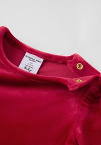 Vaaleanpunainen vauvan vaate, jossa on kultaiset nepparit kaula-aukossa ja näkyvä valkoinen etiketti, joka näyttää brändin ja koon tiedot.