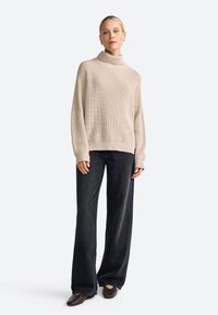 Cremfarbener Strickrollkragenpullover mit gerippter Textur, kombiniert mit dunkelblauen, weiten Jeans und braunen Slippern.