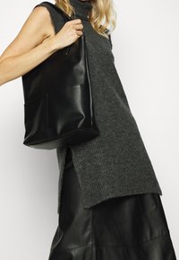 Sac cabas en cuir noir à finition brillante, présentant une forme rectangulaire, un design minimaliste et une poche avant.