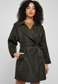 Palton trench negru, ușor, cu brâu, având o siluetă lungă, guler lat și buzunare laterale; textură netedă cu un ușor luciu.