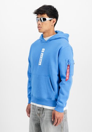 Blauwe hoodie met een voorzak, opvallend "ALPHA" logo en een met rits afsluitbare mouwzak met een rode tag. Katoenmix materiaal, casual pasvorm.