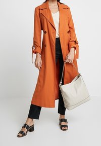 Orange trench coat med dubbelknäppning, matchad med en krämfärgad topp, mörka jeans, svarta slidesandaler och en ljusgrå handväska.