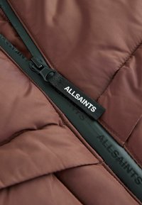 Brun pufferjacka av tyg med quiltad textur, med en svart dragkedja och ett märkesdragband med texten "ALLSAINTS."