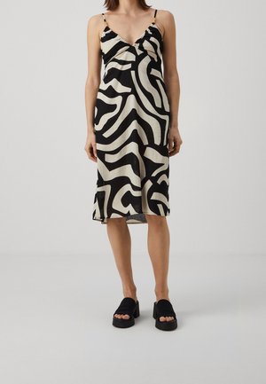 Femme portant une robe midi noire et blanche à motif abstrait sans manches, avec des fines bretelles et des sandales compensées noires.