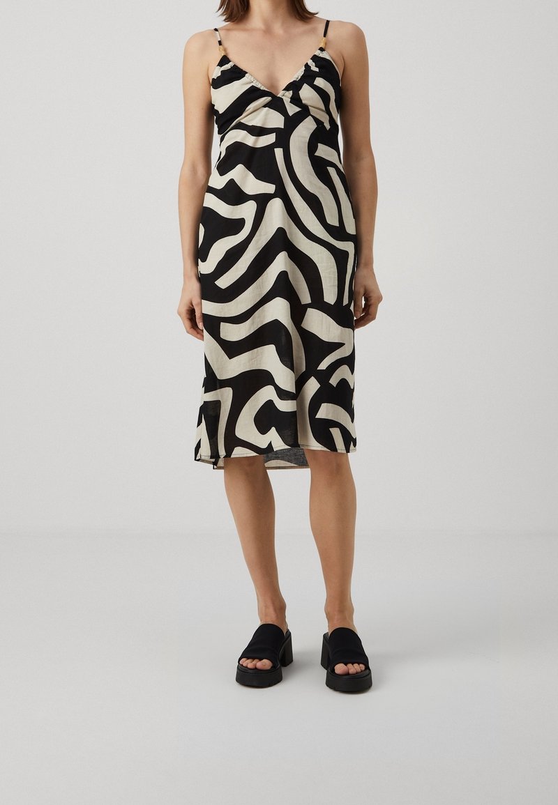 Femme portant une robe midi noire et blanche à motif abstrait sans manches, avec des fines bretelles et des sandales compensées noires.