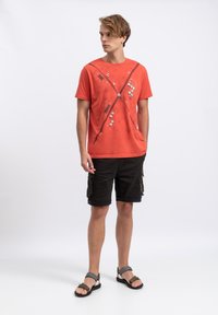 Rode T-shirt met korte mouwen en een grafisch ontwerp met lijnen en stippen, gecombineerd met zwarte cargo shorts en grijze sandalen met verstelbare banden.