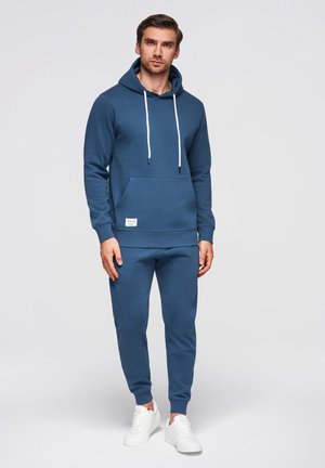 Uomo in piedi, indossa una felpa con cappuccio blu e pantaloni da jogging abbinati con scarpe da ginnastica bianche, rivolto in avanti contro uno sfondo semplice.