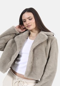 Beżowa, puszysta kurtka o kroju cropped z szerokim kołnierzem, długimi rękawami i miękką fakturą, noszona na białej dopasowanej bluzce.