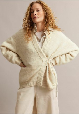 Vrouw met krullend haar, gekleed in een crèmekleurige wikkelcardigan over een overhemd met kraag en losse crèmekleurige broek, staand met handen in de zij.