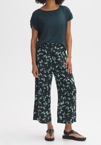 Tmavě zelená blůza s krátkými ohrnutými rukávy, v kombinaci se širokými černými kalhotami s abstraktním vzorem v mentolově zelené barvě. Černé sandály na ploché podrážce.