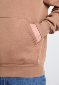 Haut à capuche marron avec poche kangourou, poignets et ourlet côtelés. Le tissu est doux avec une texture lisse, associé à un jean bleu.