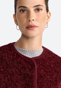 Gros plan sur une femme aux cheveux bruns, portant une veste texturée bordeaux sur un pull gris à côtes, mettant en valeur son maquillage naturel et ses boucles d'oreilles créoles.