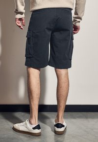 Černé cargo kraťasy s bočními kapsami, vyrobené z bavlny. Nohou v bílé tenisce s černými akcenty, stojící na šedé podlaze.