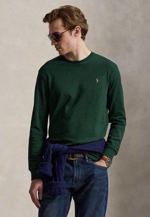 Uomo con camicia verde scuro a maniche lunghe, jeans blu, occhiali da sole, con maglione blu legato in vita, guarda a sinistra.