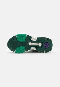 Suola di scarpa sportiva verde e nera con il sistema "TORSION" visibile e dettaglio circolare viola vicino al tallone su sfondo bianco.