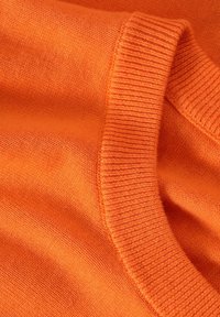 Jasno pomarańczowa dzianina o gładkiej fakturze, z prążkowanym okrągłym dekoltem i miękkim układem. Brak widocznych wzorów ani ozdób.