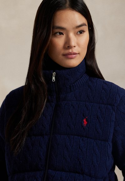Marinblå kabelstickad zip-jacka med hög krage och röd broderad logotyp på bröstet. Har en kviltad textur och robust blixtlås.
