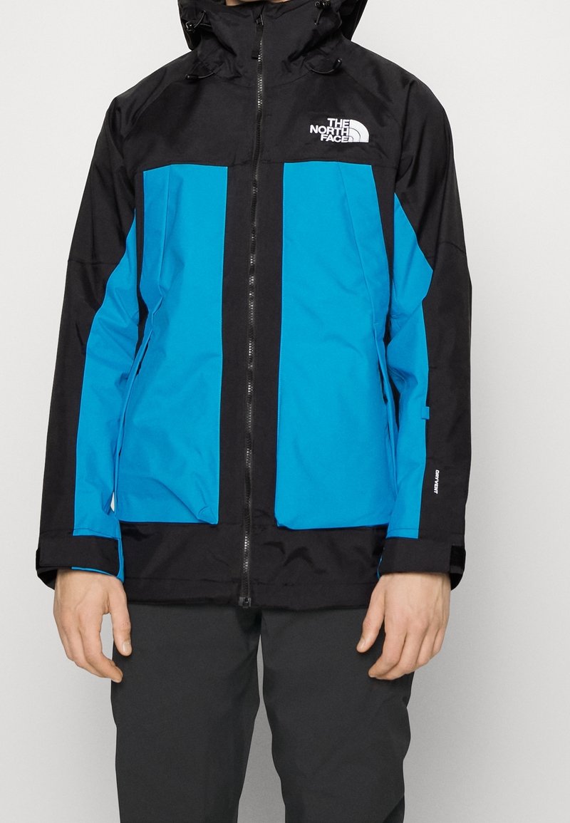Homme portant une veste imperméable North Face noire et bleue avec une fermeture éclair à l'avant et un logo sur la poitrine, debout devant un fond uni.