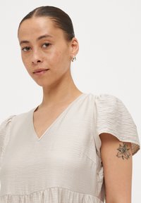 Απαλό μπεζ φόρεμα με V ντεκολτέ, φουσκωτά μανίκια, υφασμάτινη υφή και μαζεμένη μέση. Φέρει ασημένια κρεμαστά σκουλαρίκια και λουλούδι τατουάζ στο μπράτσο.