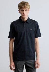 Tengerészkék pamut poloing puha tapintású anyagból, gallérral, három gombbal és egy kerek logóval a bal mellrészen.