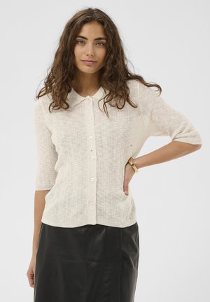 Donna con capelli lunghi e ricci che indossa un maglione beige con bottoni e maniche a gomito, e una gonna di pelle nera, con la mano sul fianco.