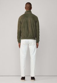 Veste bomber en suède verte avec des poignets côtelés, détail au dos, portée sur un pantalon blanc et des chaussures brunes, vue de dos.