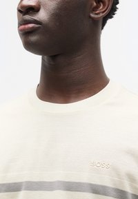 Μπεζ μάλλινο πουλόβερ με κοντά μανίκια και ριμπ υφή, με γκρι οριζόντια ρίγα και ανάγλυφο λογότυπο "BOSS" στο στήθος.