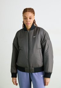 Nike bomber jakna u tamnosivoj boji, sa patentnim zatvaračem sprijeda, rebrastim ovratnikom i manžetama, uz suptilan logo na prsima. Glatka, podložena tekstura.