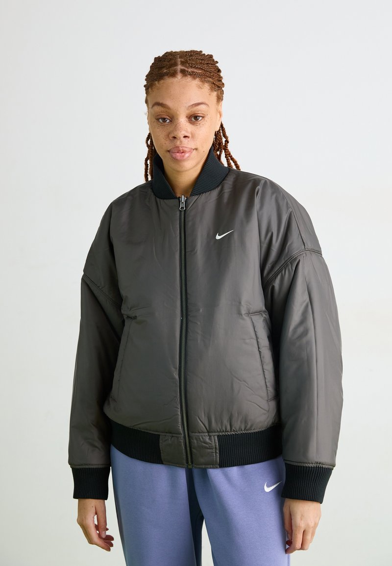 Veste bomber Nike en gris foncé, avec une fermeture éclair à l'avant, un col et des poignets côtelés, et un logo discret sur la poitrine. Texture lisse et rembourrée.